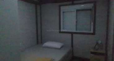 Das Bild zeigt ein kleines, schwach beleuchtetes Schlafzimmer mit einem Einzelbett, einem Kissen, einem gelben Handtuch, einem Fenster mit Fensterläden und einem Nachttisch, auf dem ein kleiner Ventilator steht.