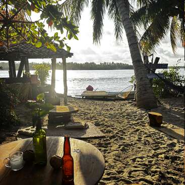 Eine ruhige Strandszene mit einem Holztisch mit Getränken, sandigem Boden, Palmen und Sitzgelegenheiten mit Blick auf einen ruhigen Fluss, über den Sonnenlicht scheint. Entspannte Atmosphäre.