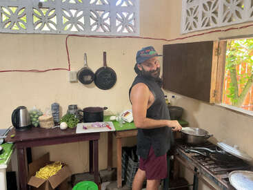 Un homme barbu, vêtu d'une chemise sans manches et d'un chapeau, est debout dans une cuisine rustique, entouré d'ingrédients, d'ustensiles et d'une fenêtre ouverte, en train de cuisiner.