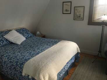 This cozy bedroom features a blue-patterned bedspread, white blanket, wooden nightstand, standing lamp, two wall art pieces, a window, and soft natural lighting.