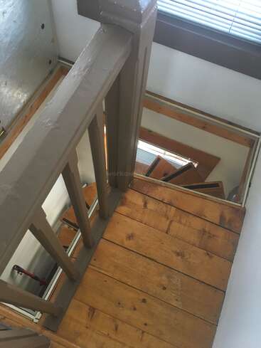 This image shows a wooden staircase with a brown railing, viewed from the top floor. Sunlight filters through blinds, casting light on the bright wooden steps.