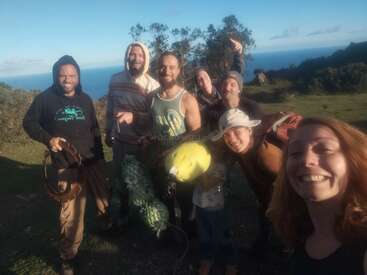 A group of seven smiling friends pose outdoors on a grassy hill with the ocean in the background, holding paragliding or adventure gear, enjoying sunny weather.