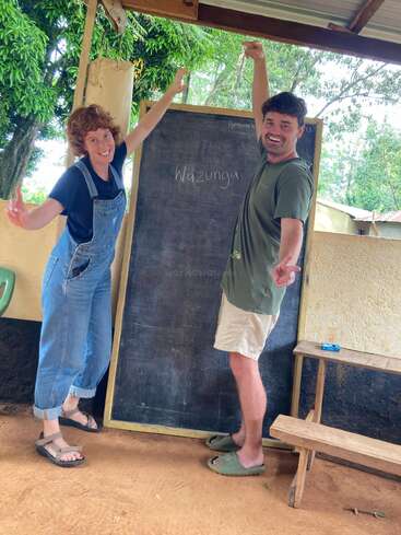 Deux personnes souriantes posent de manière ludique à côté d'un tableau noir en plein air. Le tableau noir porte l'inscription "Wazungu". Des arbres et des bancs sont visibles à l'arrière-plan, ce qui donne de la vie.
