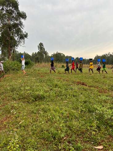 Un groupe de personnes, principalement des enfants, marchent en ligne dans un champ herbeux, chacun portant des récipients bleus sur la tête, sous un ciel couvert.