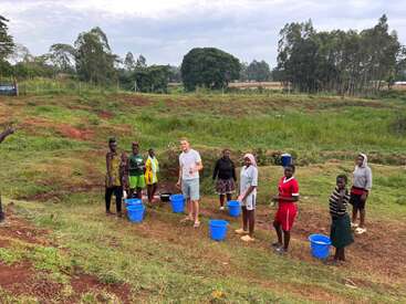 Un groupe de personnes, comprenant des enfants et des adultes, se tient en plein air sur un terrain herbeux avec des seaux bleus, probablement pour recueillir de l'eau près d'une zone rurale et verdoyante.