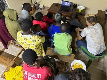 Un groupe diversifié de personnes, comprenant des enfants et des adultes, est assis sur des bancs à l'intérieur, regardant attentivement l'écran d'un ordinateur portable. Une personne mange dans un bol.