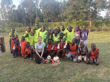 Un groupe d'enfants souriants portant des dossards orange et jaunes, ainsi qu'un adulte, posent sur un terrain herbeux avec des ballons de football, des arbres et la lumière du soleil.