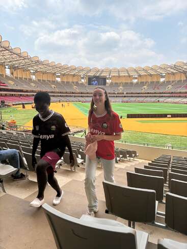 Deux personnes portant des maillots du Kenya montent les marches du stade. Le grand stade moderne est en grande partie vide, avec un terrain vert et un ciel partiellement nuageux au-dessus.