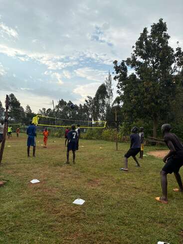 Eine Gruppe von Jungen spielt draußen auf einer Wiese, umgeben von Bäumen und unter einem teilweise bewölkten Himmel, ein energiegeladenes und lebendiges Volleyballspiel.