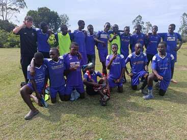 Um time de futebol de homens jovens, vestidos com uniformes azuis, posa junto em um campo gramado, alguns ajoelhados, outros em pé, sorrindo e fazendo gestos.