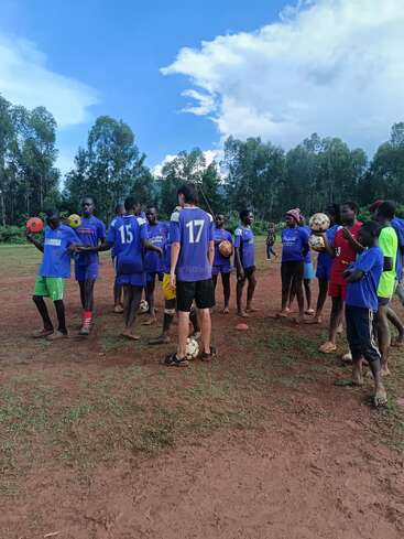 Um grupo de crianças e adolescentes, a maioria com camisetas azuis, está em um campo de futebol enlameado segurando bolas de futebol, cercado por árvores altas sob um céu parcialmente nublado.