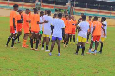 A group of young soccer players huddle together on a grassy field, wearing orange and white uniforms, seemingly preparing or strategizing before their match begins.