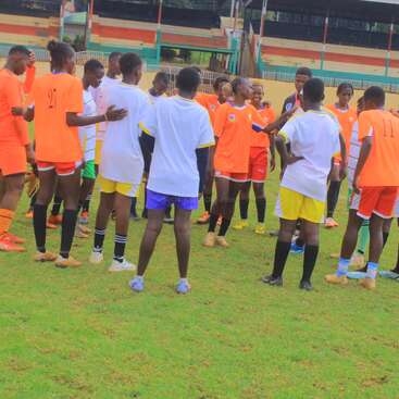 A group of young soccer players huddle together on a grassy field, wearing orange and white uniforms, seemingly preparing or strategizing before their match begins.