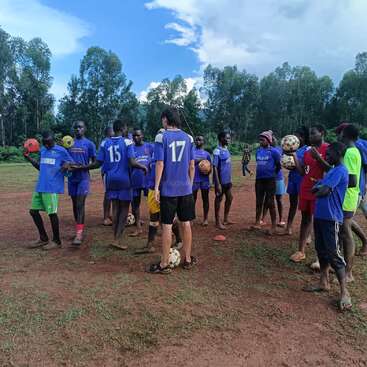 A group of children and teenagers, mostly in blue jerseys, stand on a muddy soccer field holding footballs, surrounded by tall trees under a partly cloudy sky.