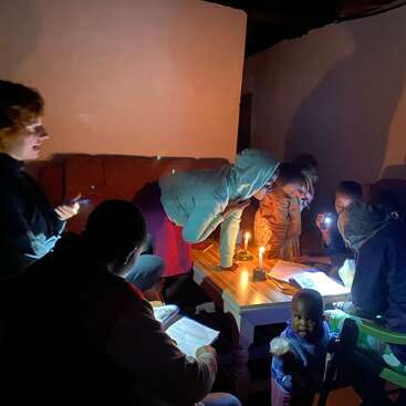 A group of people, including children, gather around a table in a dimly lit room, using candles and flashlights to read and study together.