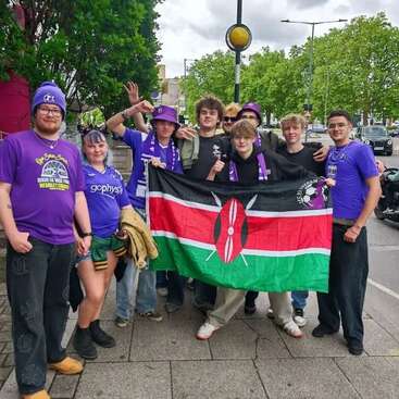 A group of young people, mostly wearing purple clothing, stand outdoors holding a Kenyan flag. They appear cheerful, posing together on a city street.