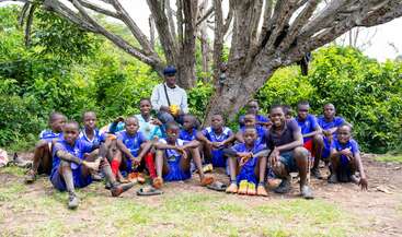 A group of young boys in blue soccer uniforms sit together under a large tree, accompanied by an adult, smiling and relaxing outdoors after a game.