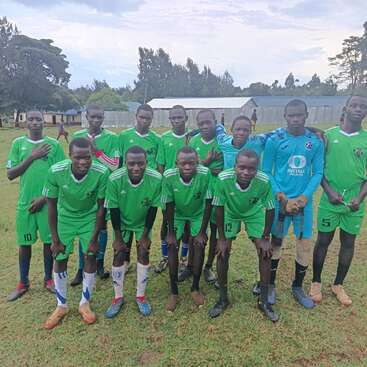 A soccer team of eleven young men in green uniforms pose together on a grassy field, with trees and buildings visible in the background, looking cheerful.