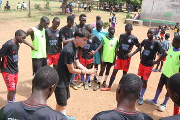 A soccer coach passionately instructs a group of teenage players gathered in a circle on a dusty field, while others watch and relax in the background.