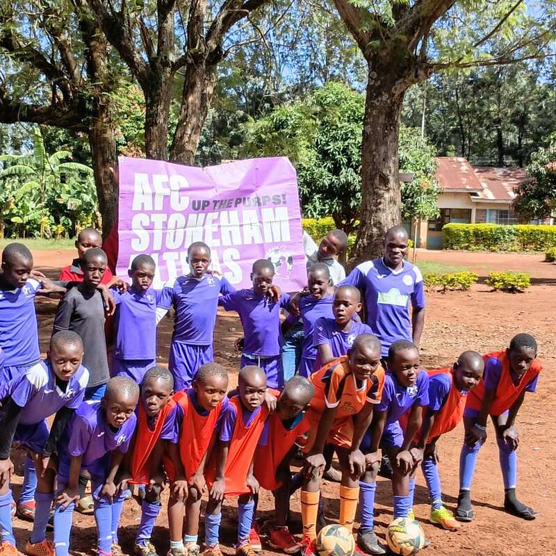 Um grupo de crianças vestindo uniformes azuis de futebol, algumas com coletes laranja, posa ao ar livre com duas bolas de futebol. Dois adultos estão atrás delas, sorrindo.