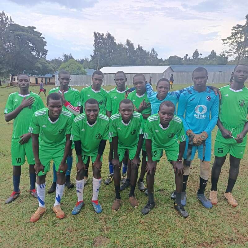 A soccer team of eleven young men in green uniforms pose together on a grassy field, with trees and buildings visible in the background, looking cheerful.