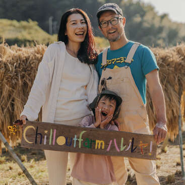 Une joyeuse famille de trois personnes se tient debout dans une ferme, avec un sourire radieux et un panneau coloré "Chillout Farm", entourée de foin récolté et de nature.