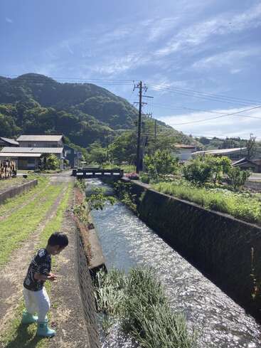 The image depicts a serene scene of a young boy standing beside a canal, surrounded by lush greenery and a bridge in the background, set against a picturesque mountainous backdrop.