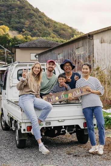 Fünf lächelnde Menschen posieren gemeinsam auf und um einen kleinen weißen Lieferwagen herum und halten ein buntes „Chilloutfarm“-Schild in den Händen. Im Hintergrund sind Berge und rustikale Gebäude zu sehen.