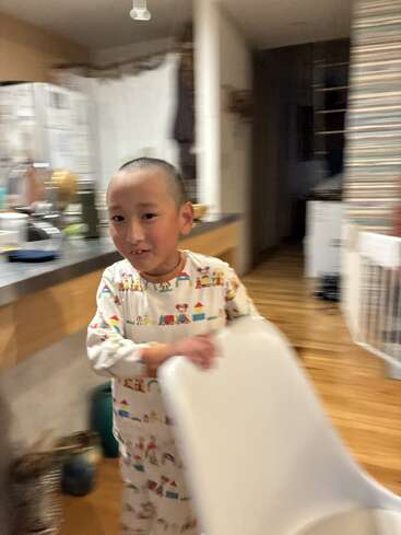 A young child with short hair smiles in a cozy kitchen, wearing playful pajamas, while holding a white chair. The background is slightly blurry with warm lighting.