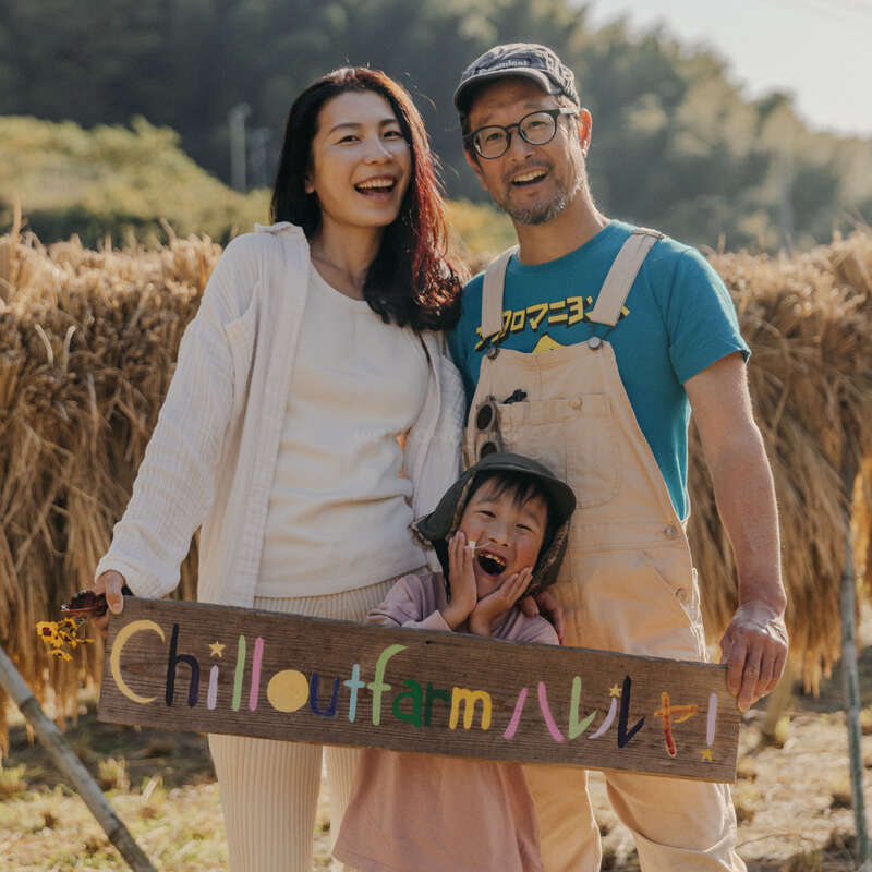 A happy family of three stands outdoors, smiling and holding a colorful "Chilloutfarm" sign. Sunflowers and golden fields create a warm, cheerful, rural atmosphere.