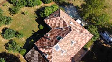 This aerial image shows a house with a red-tiled roof surrounded by trees and greenery. A driveway and several vehicles are visible near the building.