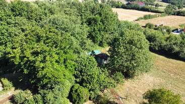This aerial image shows a small, rustic cabin surrounded by dense green trees. Open fields and other vegetation are visible in the background under bright sunlight.