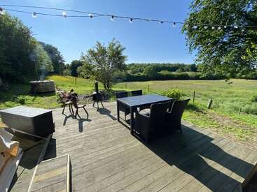 A wooden deck overlooks a serene green field and forest. Outdoor furniture, string lights, and a fire pit create a peaceful, inviting atmosphere under the clear blue sky.