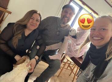 A group of four people, including a child with an emoji face, sit smiling indoors with a white dog. Everyone appears happy and relaxed together.