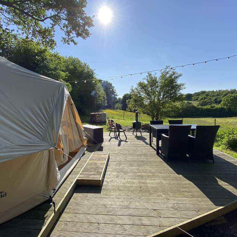 A cozy glamping tent sits on a wooden deck with outdoor furniture, under a bright sunny sky, surrounded by green trees and an open meadow. Peaceful setting.