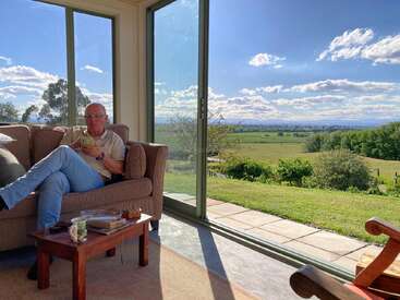A man relaxes on a sofa, using his phone, inside a sunlit room with large windows showcasing a scenic, green countryside landscape under a bright sky.