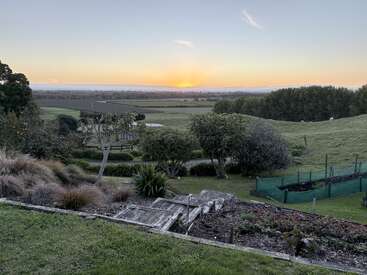 A beautiful countryside landscape at sunrise, featuring green fields, gardens, wooden steps, trees, and distant hills under a clear sky, radiating peace and tranquility.