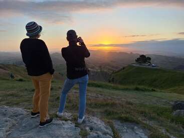 Two people stand on a rocky hilltop, watching and photographing a stunning sunset over rolling green hills, with a distant car parked near a lone tree.