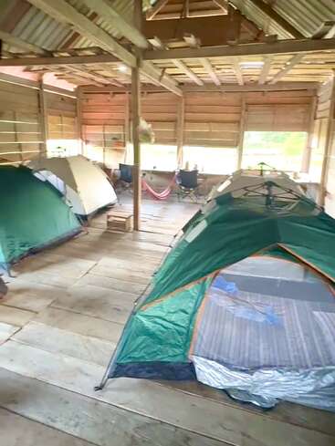 Inside a rustic wooden shelter, several tents are set up on a wooden floor. Camping chairs, a hammock, and soft daylight create a cozy, communal atmosphere.