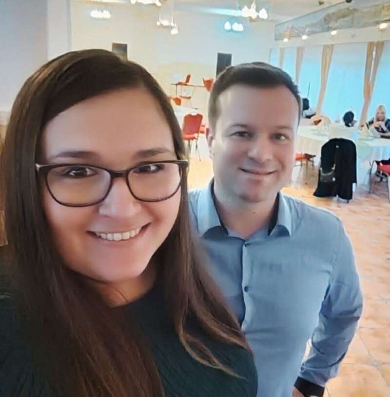 A smiling woman with long dark hair and glasses stands next to a man in a light blue shirt. They are inside a bright, spacious room.