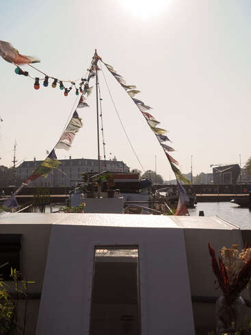 Ein mit bunten Fahnen und Glühbirnen geschmücktes Boot liegt in einem Kanal vor Anker, mit Gebäuden und Sonnenlicht im Hintergrund, was eine festliche, friedliche Szene ergibt.