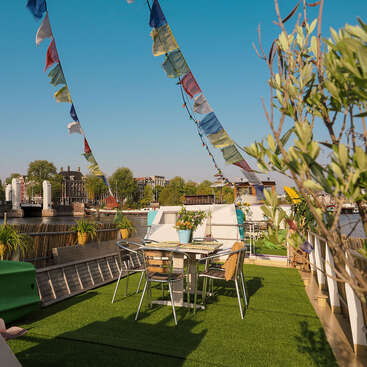 This image shows a cozy outdoor seating area on a houseboat, decorated with colorful flags, plants, and a table, overlooking a peaceful riverside cityscape.