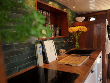 Diese gemütliche Küche hat grün geflieste Wände, hölzerne Arbeitsplatten, ein Schneidebrett, fröhliche gelbe Blumen in einer Vase, offene Regale mit Gläsern und eine warme, einladende Beleuchtung.