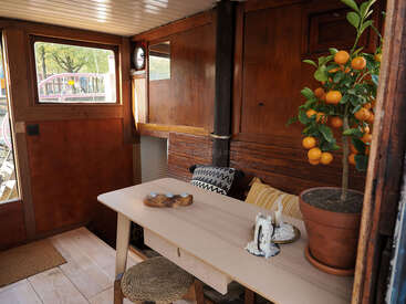 Diese gemütliche Holzeinrichtung besteht aus einem einfachen Tisch, geflochtenen Hockern und dekorativen Kissen. Ein getöpferter Zitrusbaum bringt Farbe ins Spiel, und die Fenster sorgen für natürliches Licht. Einladende Atmosphäre.