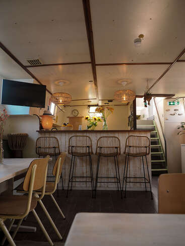 Das gemütliche Café ist mit Holzmöbeln, Barhockern aus Rattan, hängenden Korblampen, frischen Sonnenblumen, einem Fernseher und einer Treppe ausgestattet, die nach oben führt und eine warme, einladende Atmosphäre schafft.