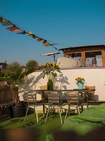 Auf einer sonnigen Dachterrasse umgeben Metallstühle einen Glastisch mit Sonnenblumen in einer Vase, Lichterketten über dem Kopf und dekorative Pflanzen schaffen eine gemütliche Atmosphäre.