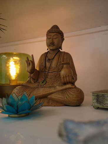 Eine ruhige hölzerne Buddha-Statue sitzt im Schneidersitz und ist mit einem Anhänger geschmückt. Umgeben von einem blauen Lotus-Kerzenhalter, einer Lampe und einer verschnörkelten Kiste, strahlt sie eine friedliche Atmosphäre aus.