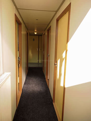 Ein schmaler Flur mit mehreren geschlossenen gelben Türen auf beiden Seiten, Teppichboden, einem Heizkörper auf der linken Seite und hellem Sonnenlicht, das von rechts hereinströmt.