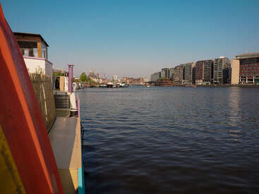Ein ruhiger Fluss fließt zwischen einem bunten Boot auf der linken Seite und modernen Gebäuden auf der rechten Seite, unter einem klaren blauen Himmel mit entfernten Stadtstrukturen.