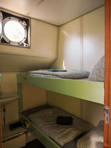 Dieses Bild zeigt ein kleines Zimmer mit Etagenbetten, gemusterter Bettwäsche, einem kleinen runden Bullaugenfenster, einem Waschbecken und minimalistischen Möbeln, die eine gemütliche, einfache Atmosphäre schaffen.